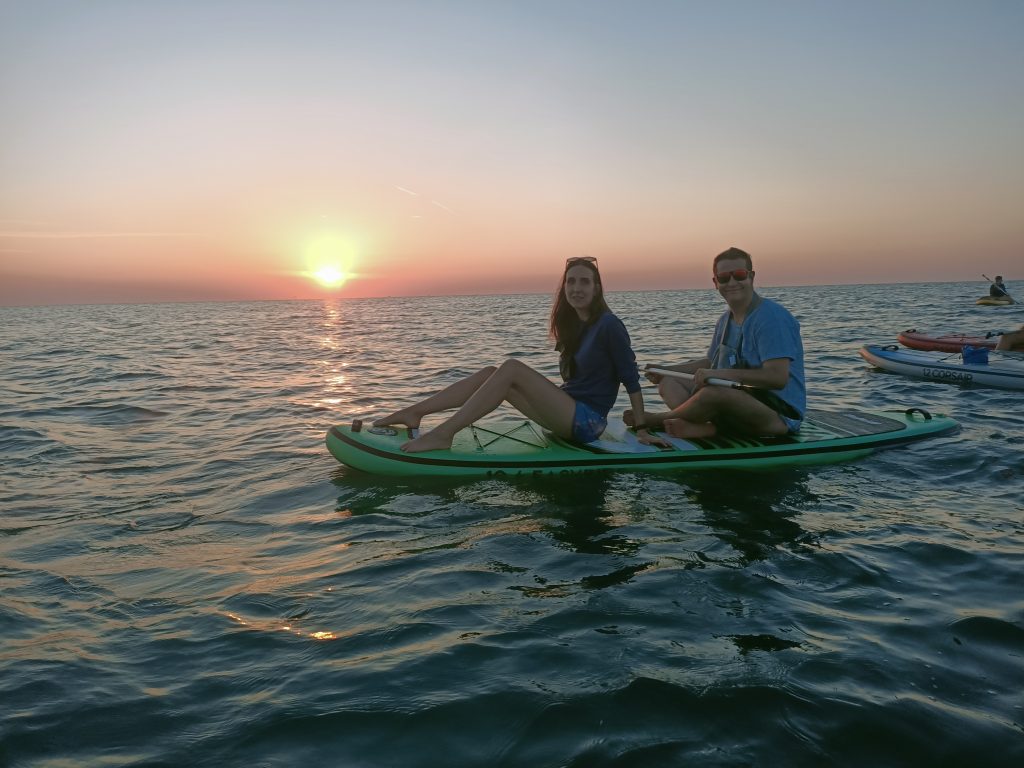 Due Giovani Soci RomagnaBanca sorridono seduti sulla tavola SUP con il sole all'orizzonte a Cesenatico
