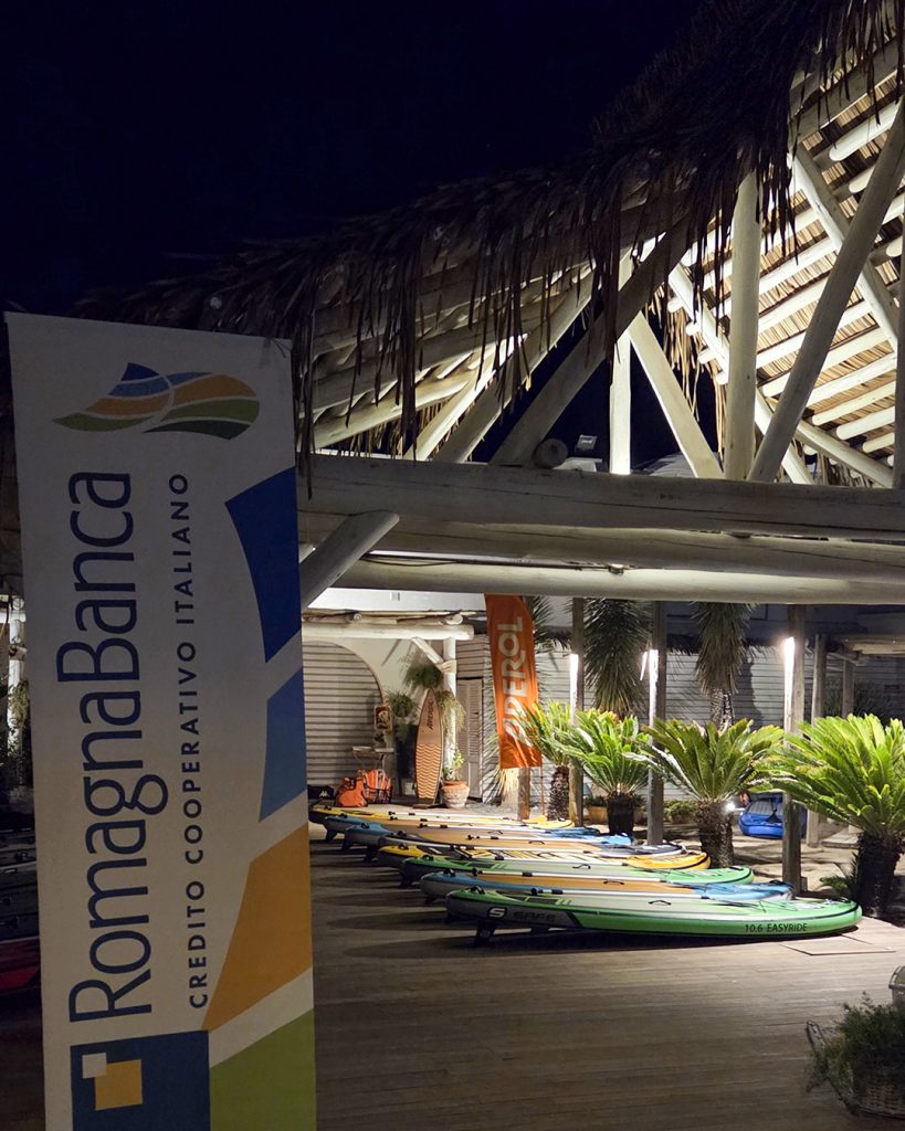 Insegna RomagnaBanca all'ingresso del Bagno Cala Romeo a Cesenatico, tavole SUP allineate prima dell'alba in Sup dei Giovani Soci