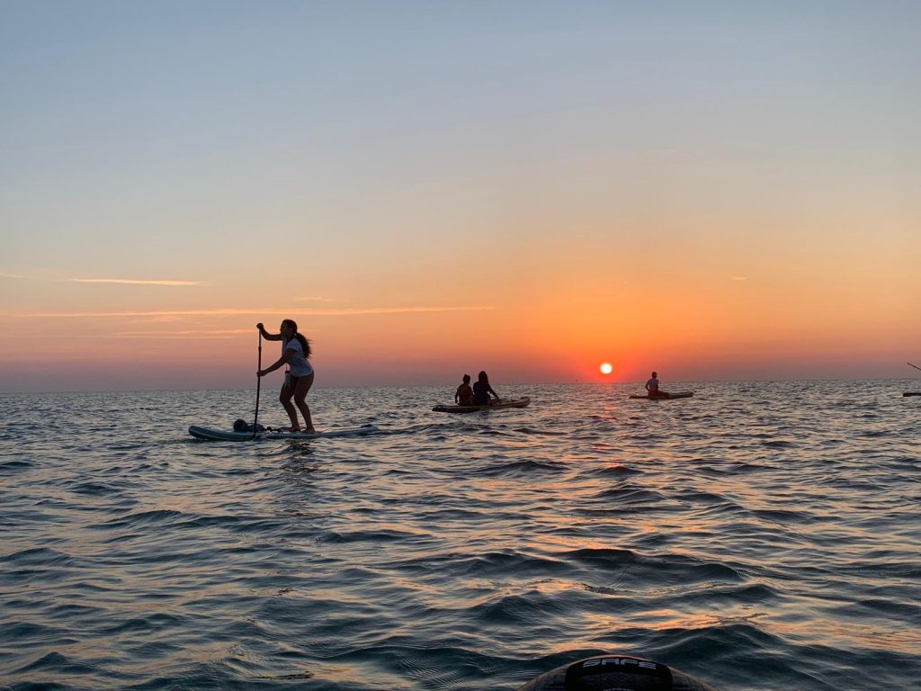 Una Giovane Socia RomagnaBanca in piedi sulla tavola SUP durante l'alba a Cesenatico, con il sole che sorge sull'orizzonte