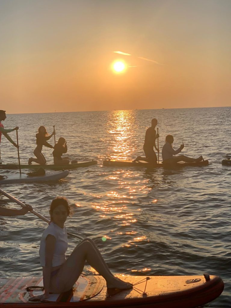 Una Giovane Socia RomagnaBanca in primo piano sulla tavola SUP, con il gruppo e il sole all'orizzonte sullo sfondo